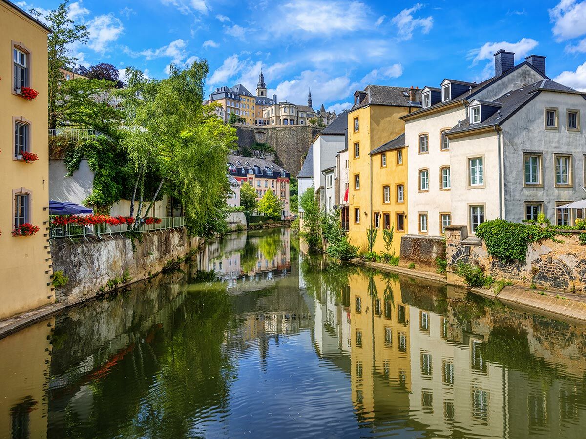 Blick auf Luxemburgs malerische Altstadt mit bunten Häusern am Fluss, grünen Bäumen und historischen Gebäuden auf dem Hügel, blauer Himmel.