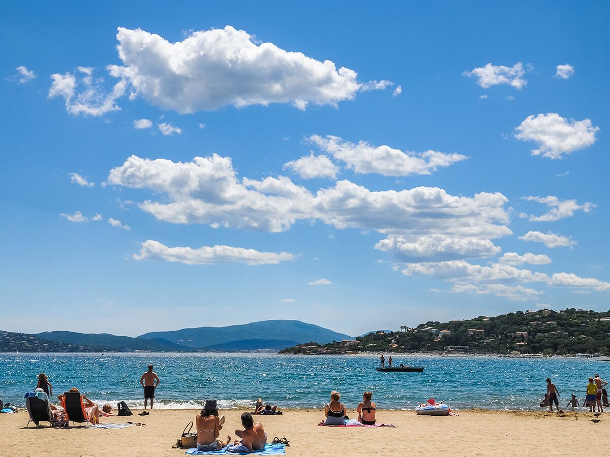 Menschen entspannen am sonnigen Sandstrand mit blauem Meer, Bergen und weißen Wolken am Himmel.