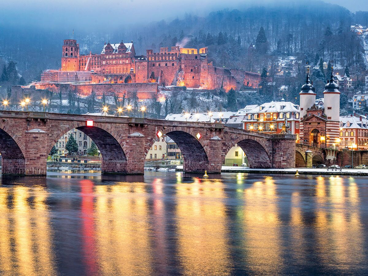 Winterabend in Heidelberg mit beleuchtetem Schloss, Alter Brücke über Neckar und Stadtlichtern im Schnee.