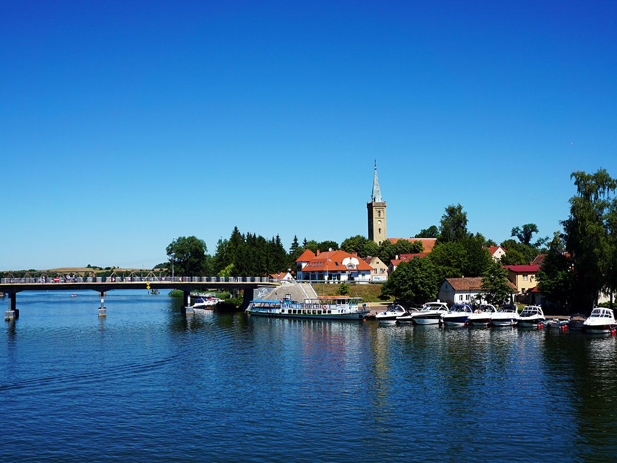 Panoramablick auf eine malerische polnische Stadt am Wasser mit Brücke, Schiffen, einem Kirchturm und viel Grün.
