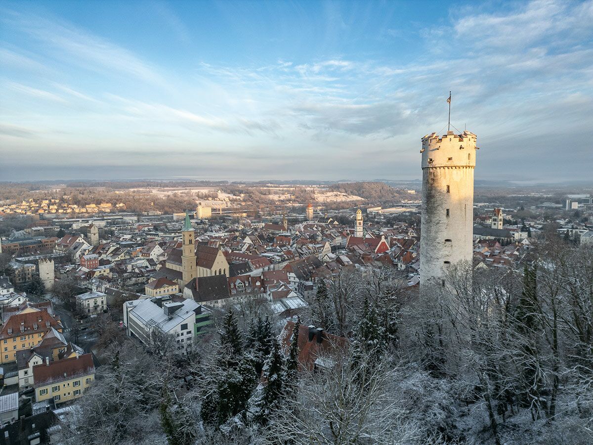 Historischer weißer Turm auf schneebedecktem Hügel über verschneiter Altstadt mit Dächern und Kirche unter blauem Winterhimmel.