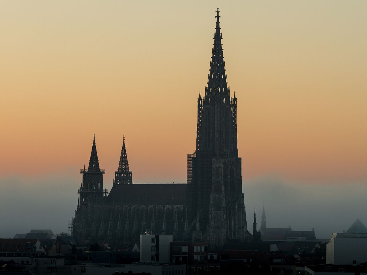Silhouette des Ulmer Münsters mit hohen Türmen bei Sonnenaufgang oder Sonnenuntergang in einer nebligen Stadtansicht.