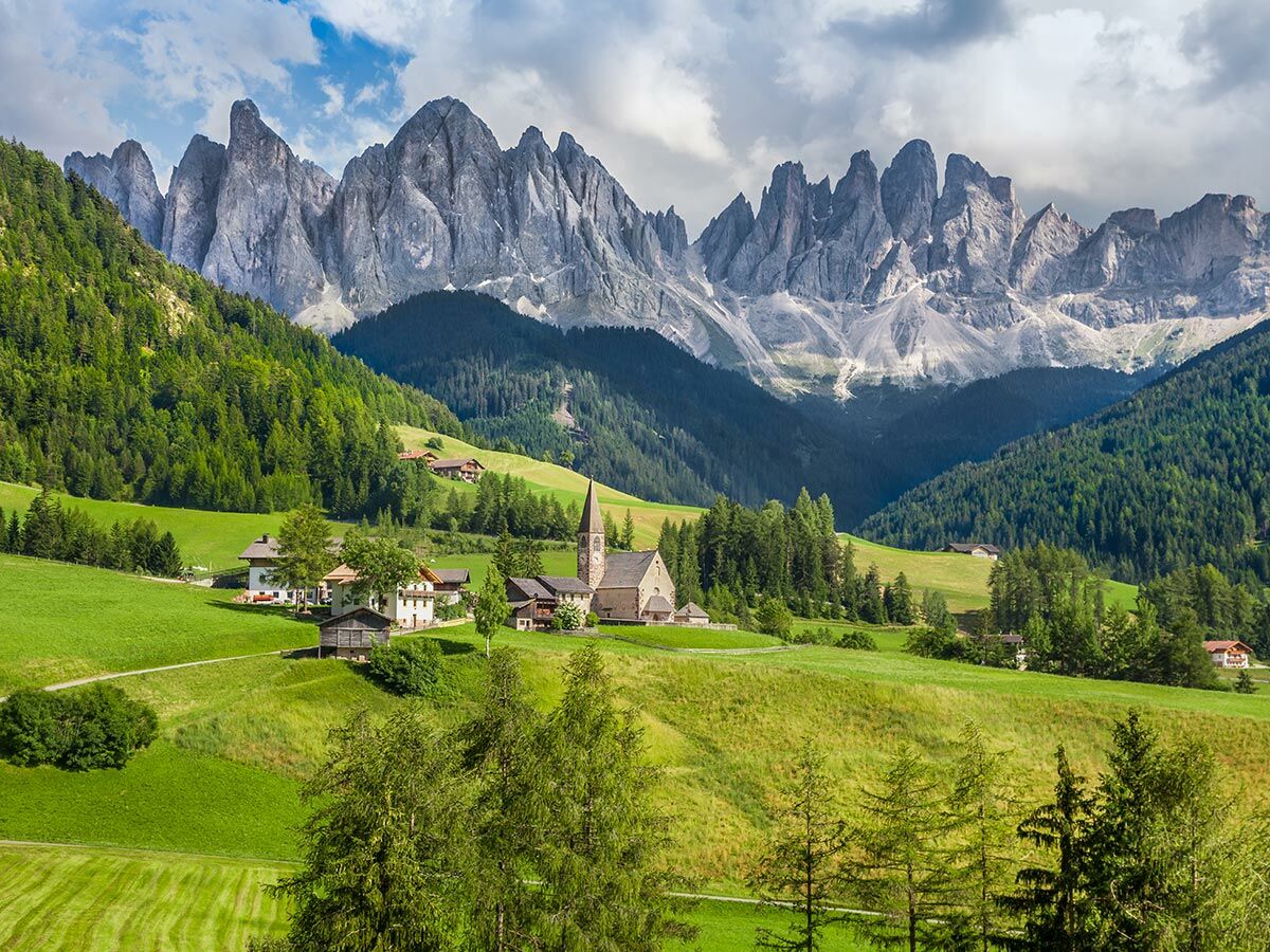 Idyllisches Bergdorf Santa Maddalena in den Dolomiten mit grünen Wiesen, Wäldern und markanten Berggipfeln unter bewölktem Himmel.