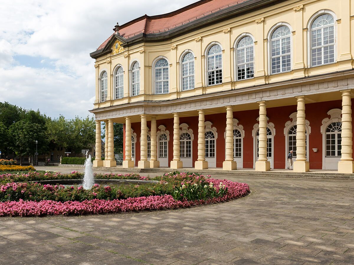 Historisches Schlossgebäude mit gelber Fassade, roten Säulen und Gartenanlage mit pinken Blumen und Springbrunnen.
