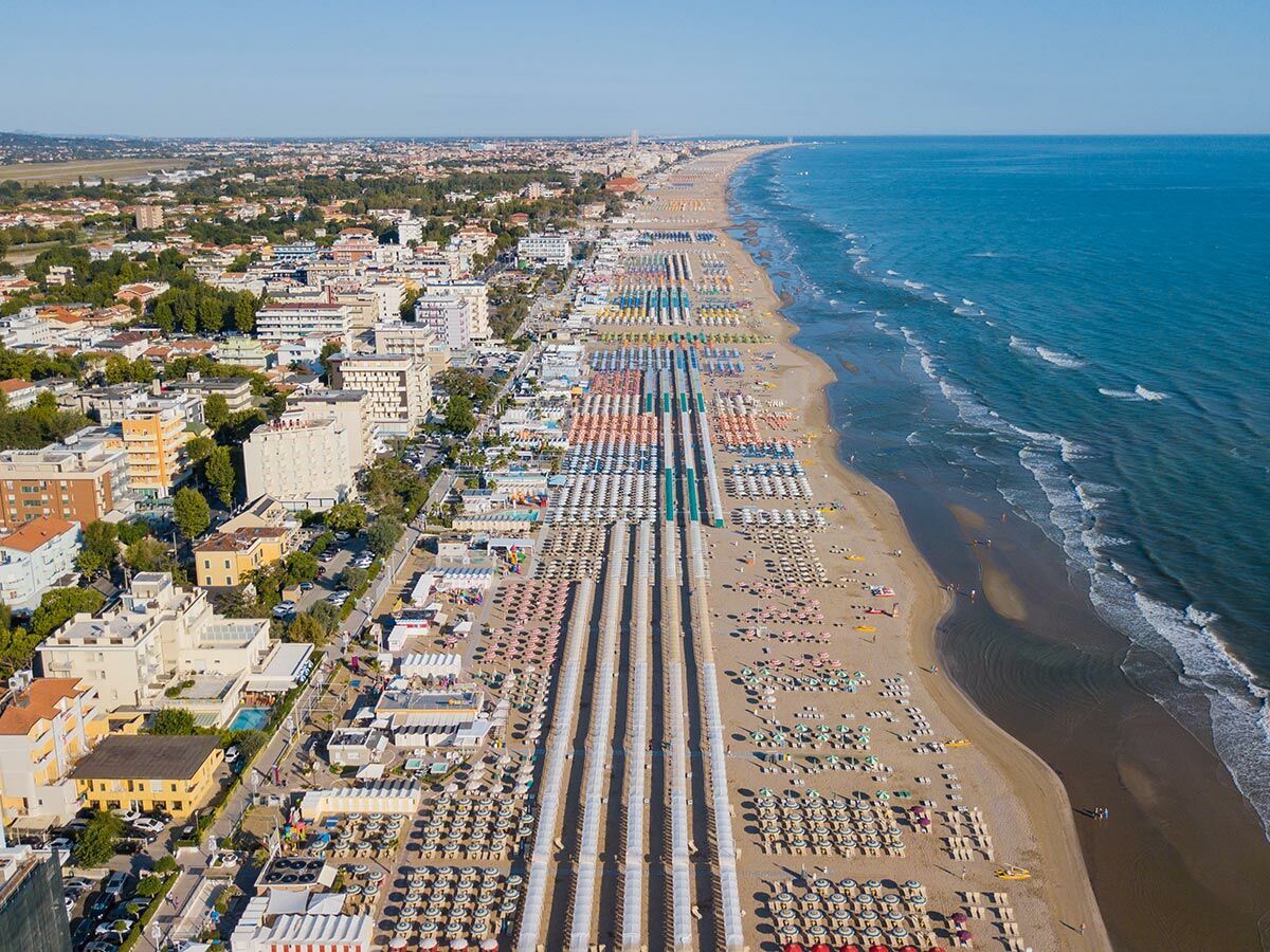 Luftaufnahme eines langen, belebten italienischen Sandstrandes mit zahlreichen bunten Sonnenschirmen, Hotels und der blauen Adria.