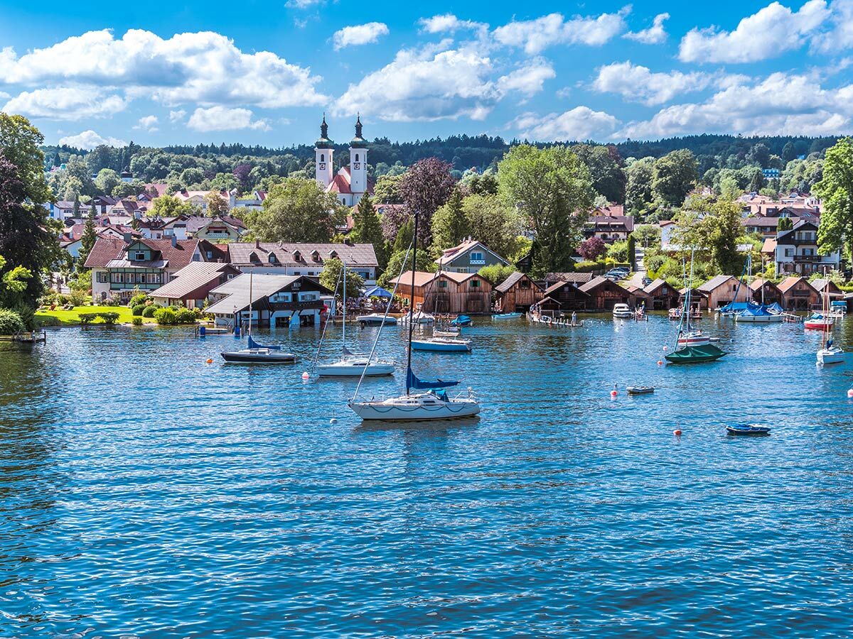 Panoramablick auf ein bayerisches Seedorf mit Kirche, Häusern und vielen Booten auf dem blauen Wasser unter hellem Himmel.
