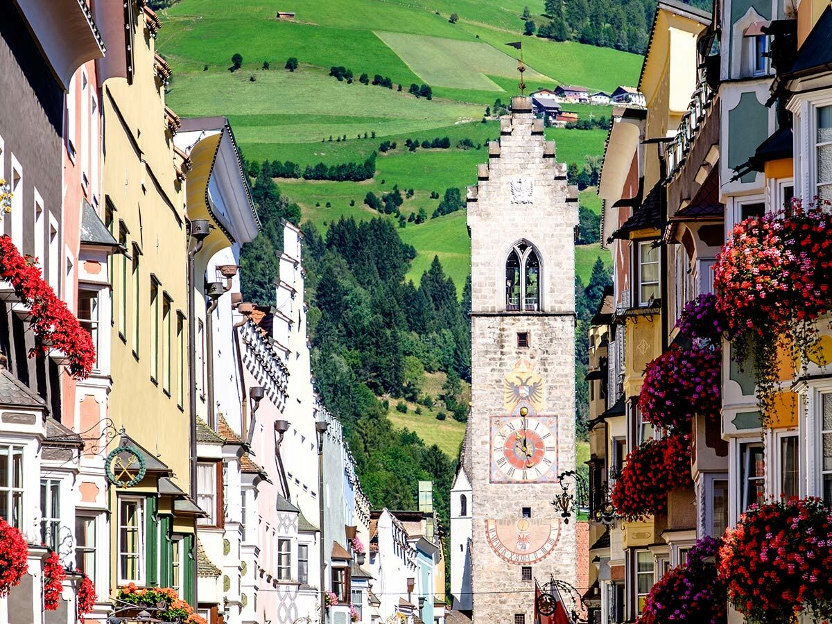 Bunte Gebäude und Blumenkästen in der historischen Altstadt von Sterzing, Südtirol, mit dem Zwölferturm und grünen Alpenhügeln im Hintergrund.