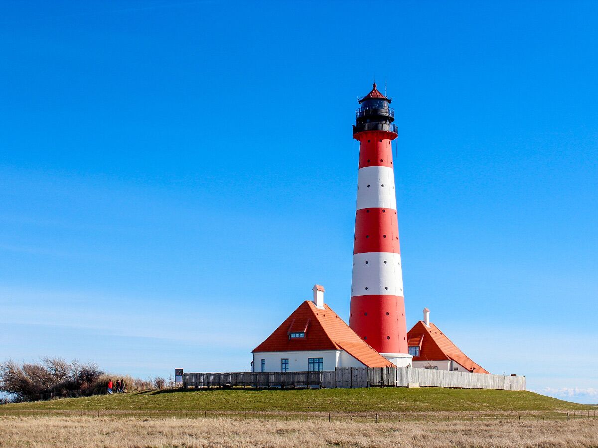 Rot-weiß gestreifter Leuchtturm Westerheversand auf grünem Hügel mit roten Häusern unter blauem Himmel an der Nordsee.