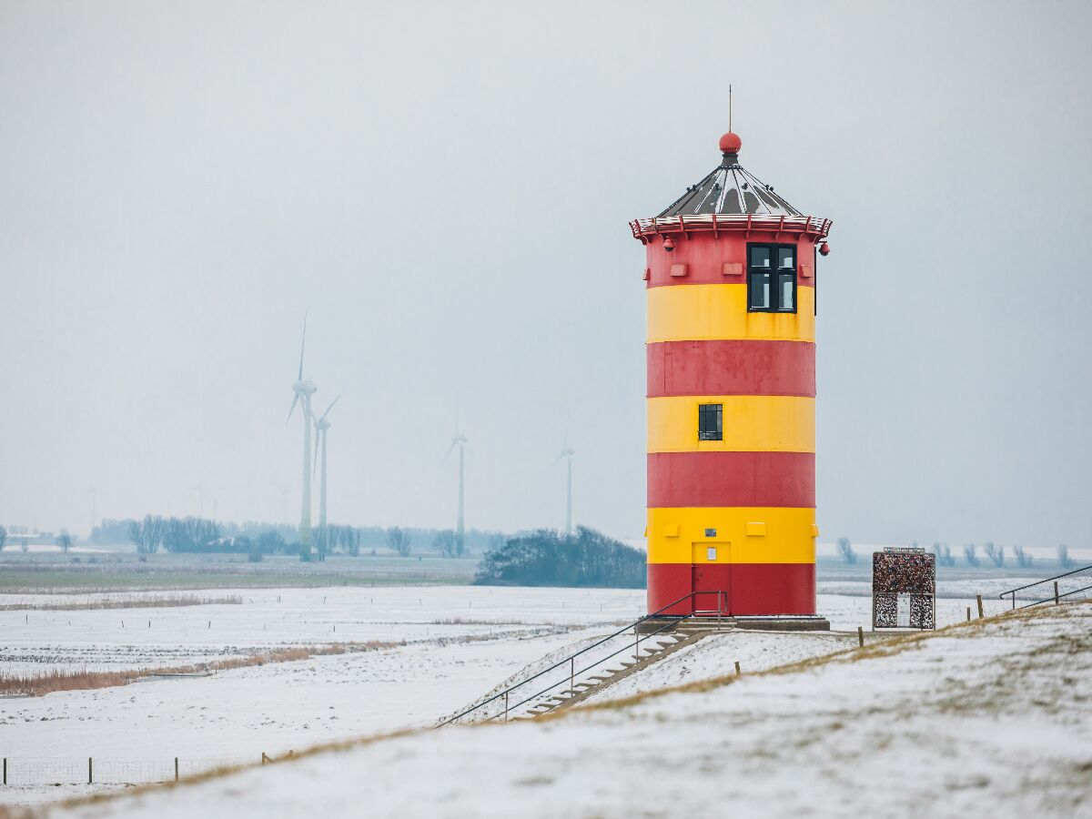 Gelb-rot gestreifter Leuchtturm steht auf einem schneebedeckten Deich vor Windrädern und grauem Winterhimmel.
