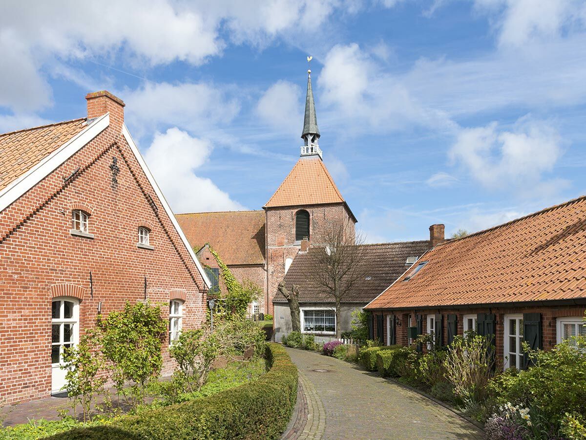 Idyllische Dorfstraße mit roten Backsteinhäusern und einer Kirche mit spitzem Turm unter blauem Himmel und weißen Wolken.
