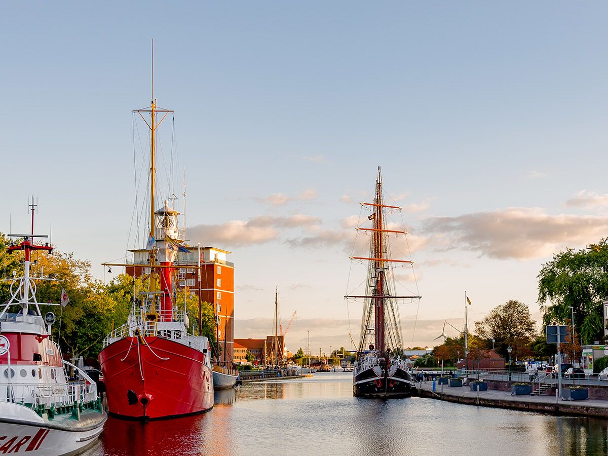 Historische Schiffe, darunter ein rotes Boot und ein Segelschiff, liegen bei Abendlicht in einem ruhigen Hafenkanal.