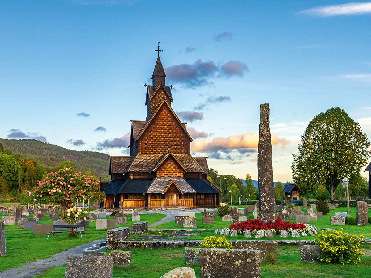 Historische Stabkirche aus Holz in Norwegen, umgeben von einem Friedhof mit alten Grabsteinen, Blumen und einem Steinmonument.