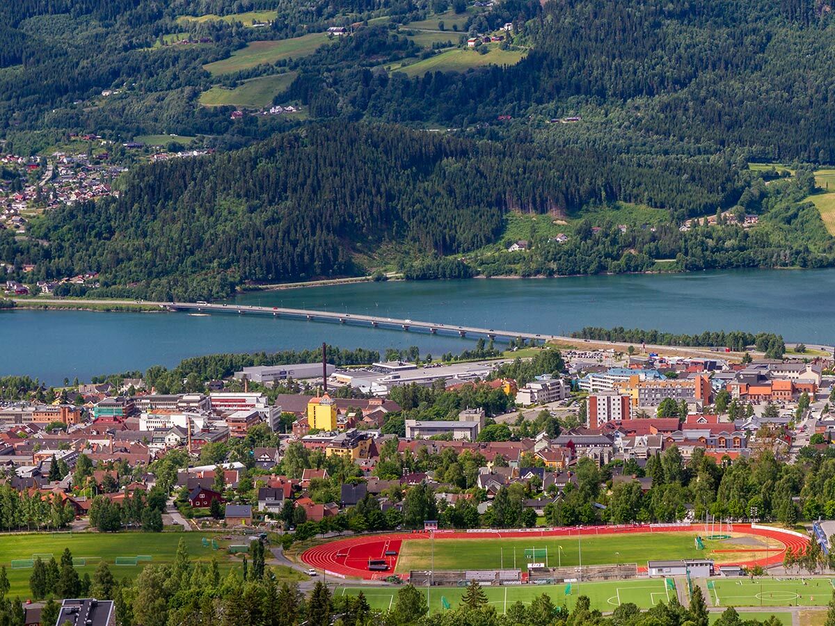 Panoramablick auf eine norwegische Stadt mit See, Brücke und Stadion, umgeben von Wäldern und Bergen.