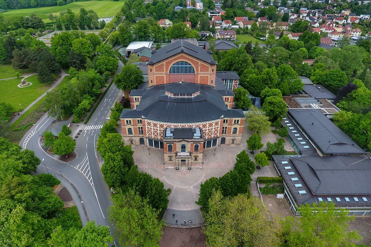 Luftaufnahme des Bayreuther Festspielhauses, umgeben von grünen Bäumen, Straßen und Wohngebäuden unter blauem Himmel.