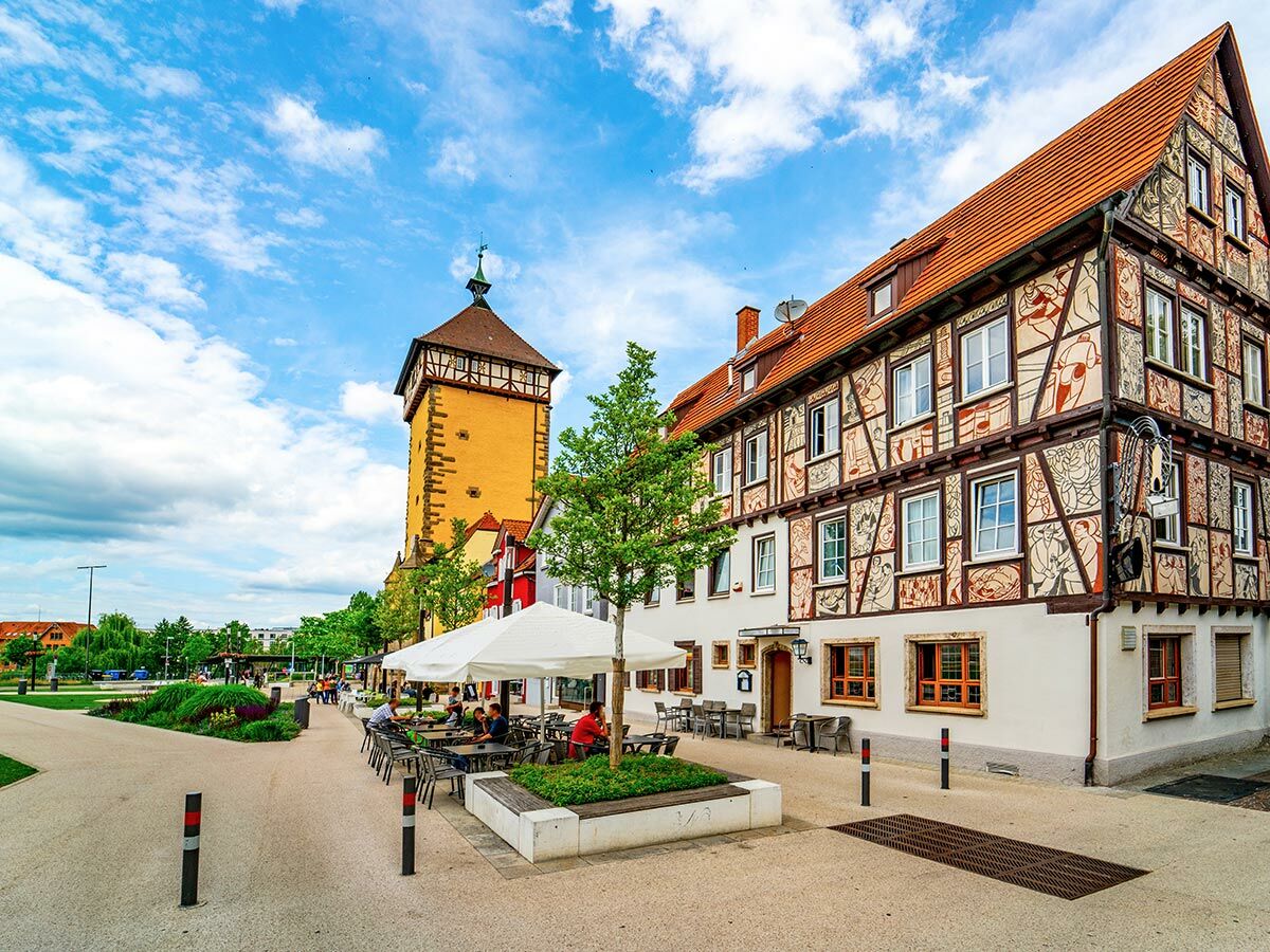 Historische Altstadt mit markantem gelbem Turm, bunt bemaltem Fachwerkhaus und belebtem Straßencafé in sonniger Fußgängerzone.