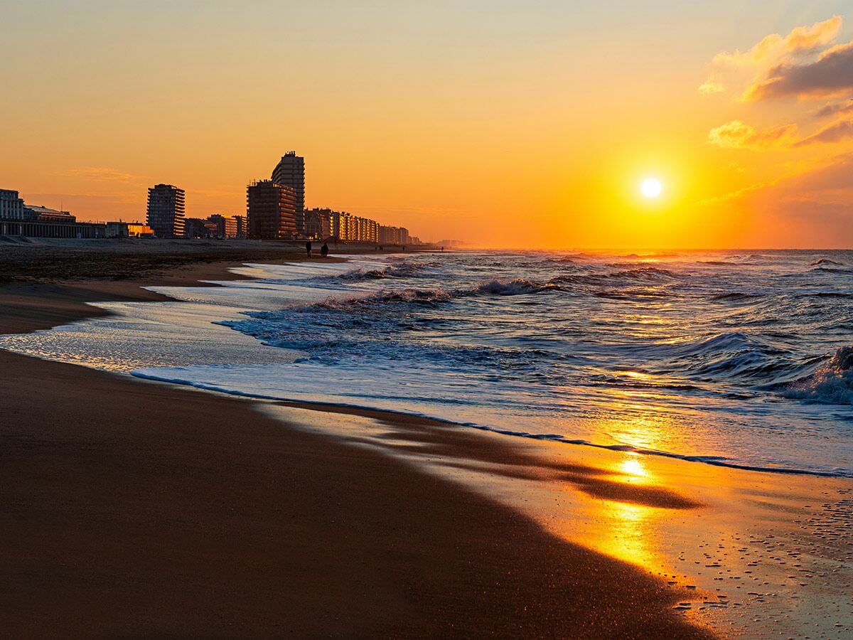Goldener Sonnenuntergang über einem belebten Stadtstrand mit sanften Wellen und der Küstenlinie im Hintergrund.
