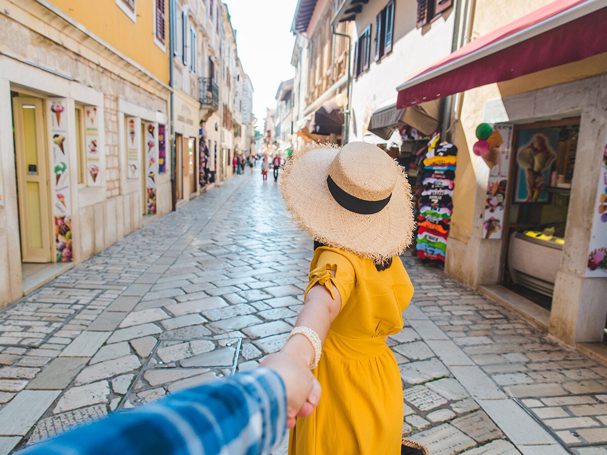 Frau in gelbem Kleid und Strohhut führt Hand in Hand durch eine sonnige, gepflasterte Altstadtgasse mit Geschäften.