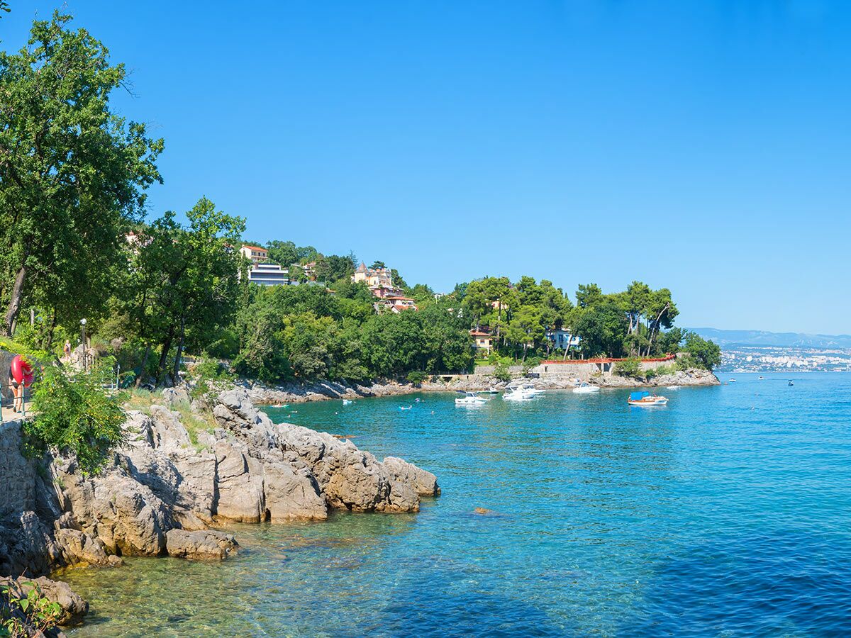 Sonnige kroatische Küste mit klarem türkisfarbenem Meer, felsiger Bucht, grünen Bäumen, Häusern auf Hügeln und Booten in der Ferne.