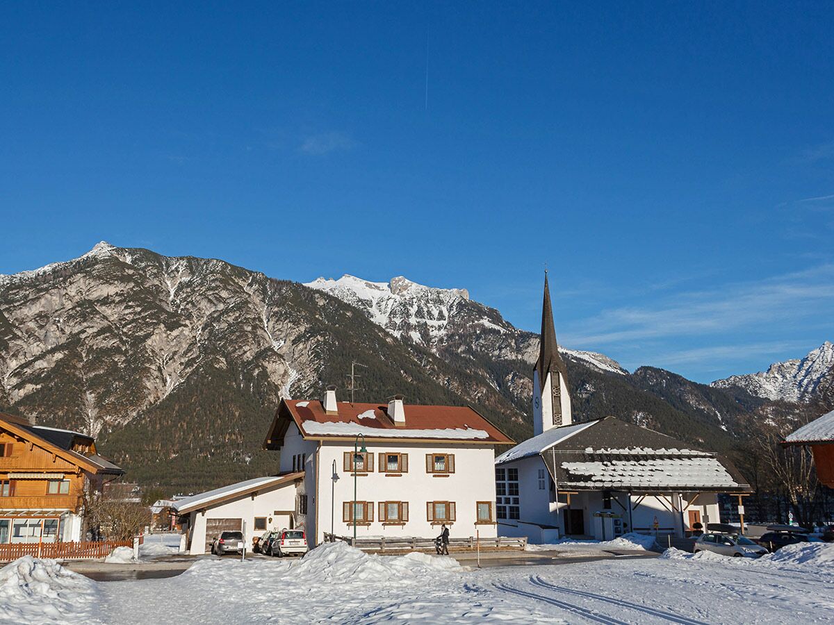 Winterliche Alpenlandschaft mit verschneitem Dorf, traditionellen Häusern, Kirchturm und majestätischen Bergen unter blauem Himmel.