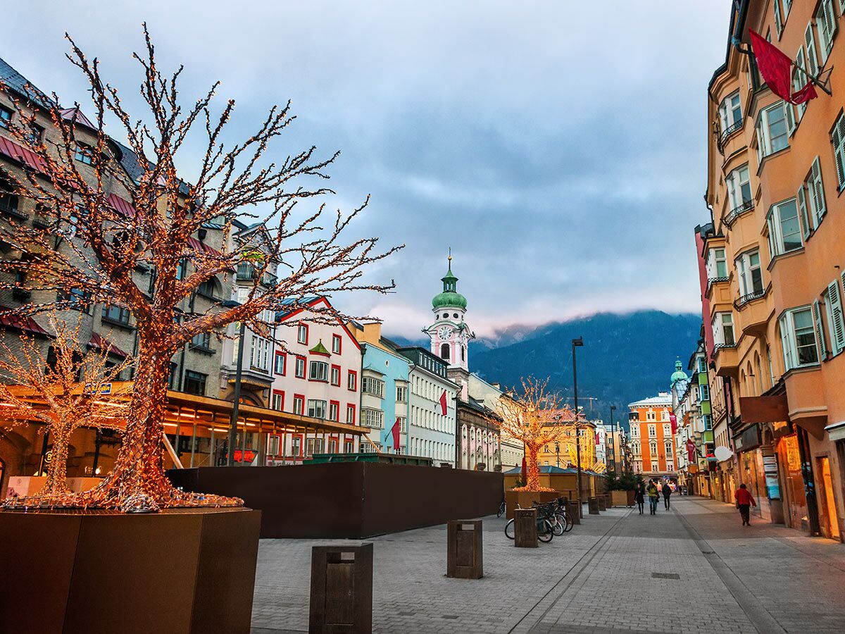 Abendliche Ansicht der Innsbrucker Altstadt mit beleuchteten Deko-Bäumen, bunten Häusern und Alpen im Hintergrund.