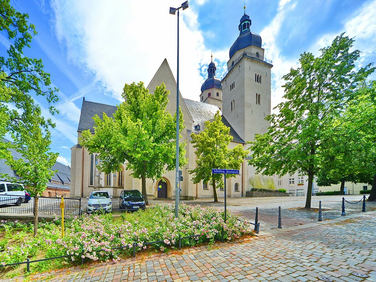 Historische Kirche mit zwei Türmen am Kopfsteinpflasterplatz in Schulberg Kirchstraße, umgeben von grünen Bäumen und Blumen.