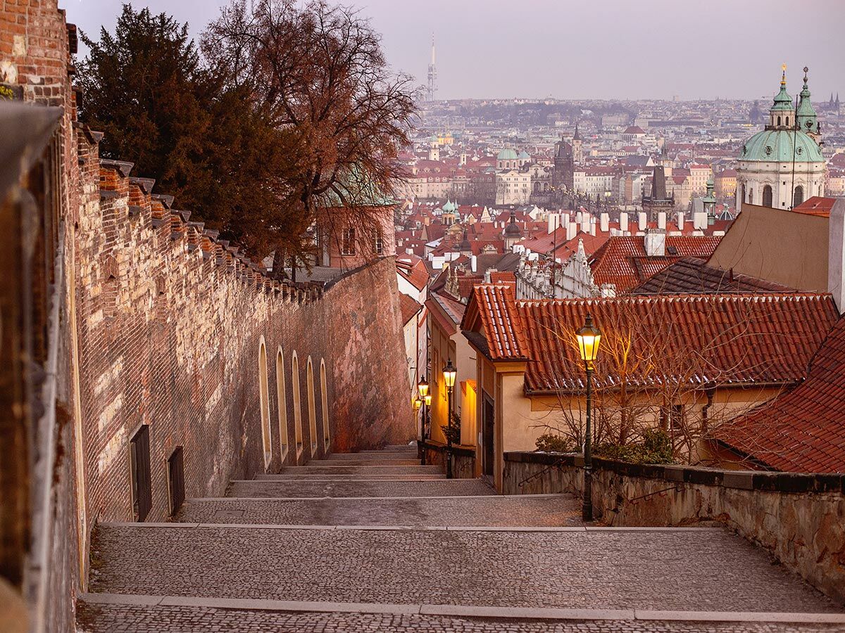 Alte Kopfsteinpflastertreppe in Prag bei Abend, beleuchtete Laternen, historische Backsteinmauer und Dächer der Altstadt.