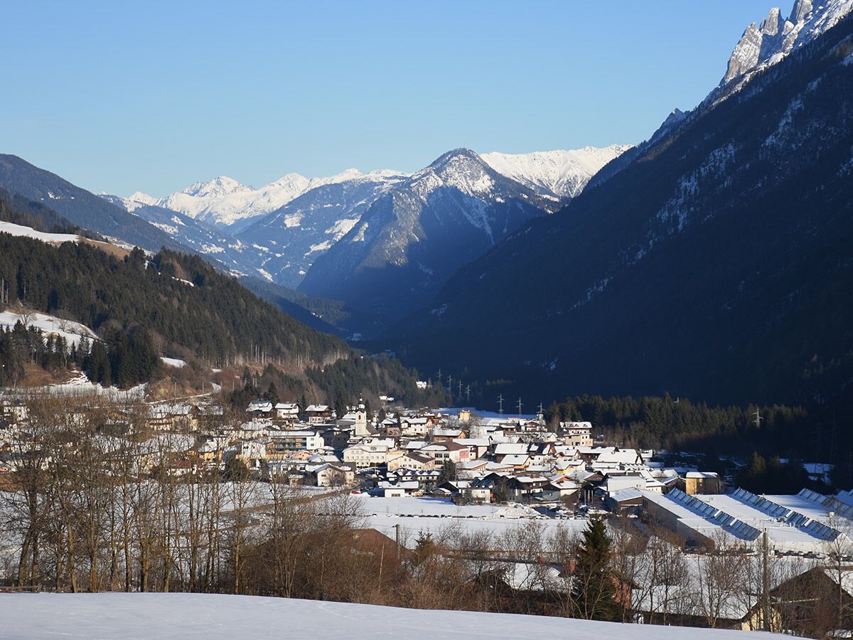 Winterlandschaft mit einem schneebedeckten Alpendorf, umgeben von bewaldeten Hügeln und majestätischen Bergen unter blauem Himmel.