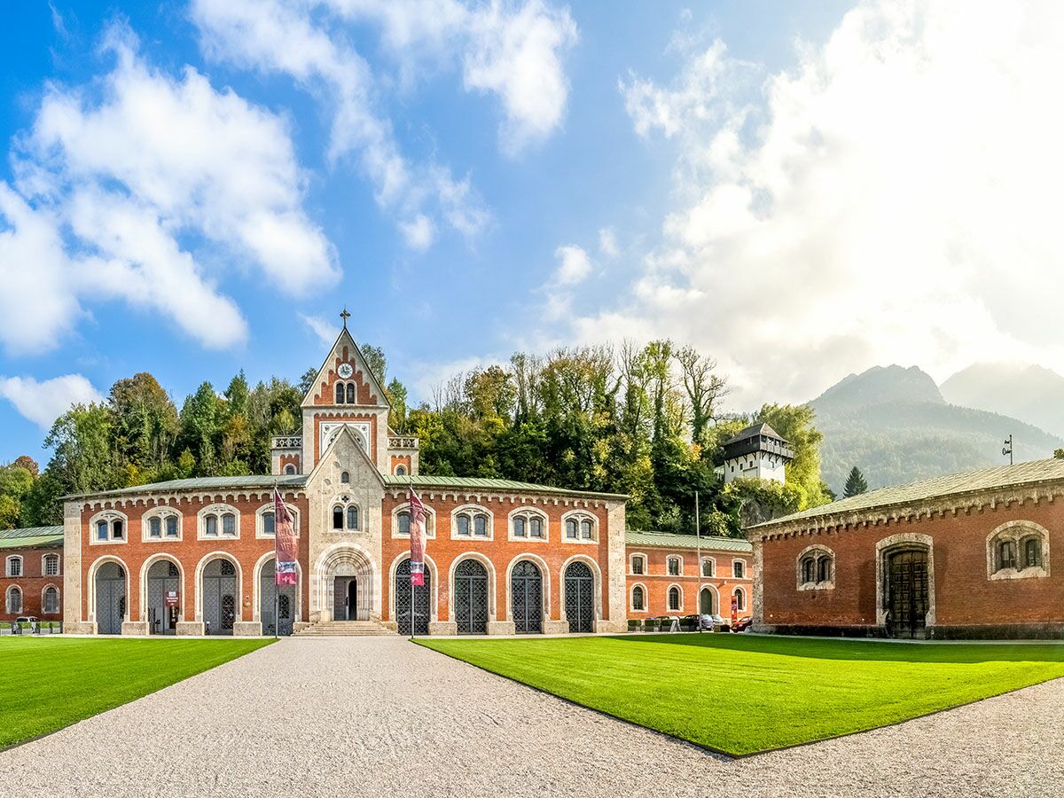 Historisches Backsteingebäude des Marstalls in Hellbrunn, Salzburg, mit Uhrturm, grünen Rasenflächen und Bergen unter blauem Himmel.