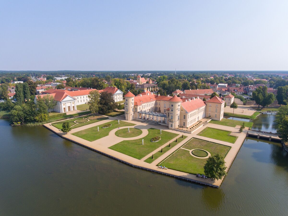 Luftaufnahme von Schloss Rheinsberg am See mit gepflegten Barockgärten und umliegender Stadt unter blauem Himmel.