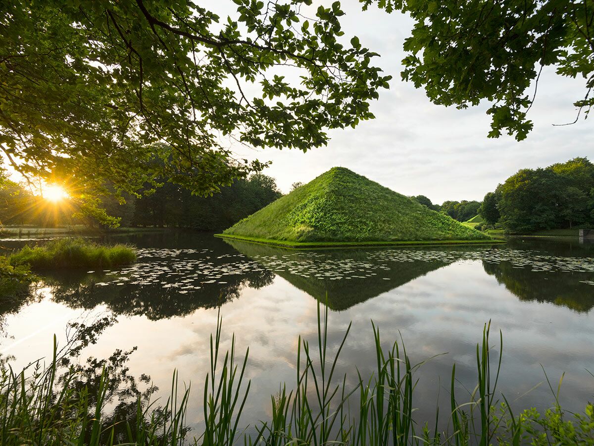 Grüne Pyramide am See mit Sonnenstrahlen bei Sonnenuntergang, gespiegelt im Wasser mit Seerosen und Bäumen.