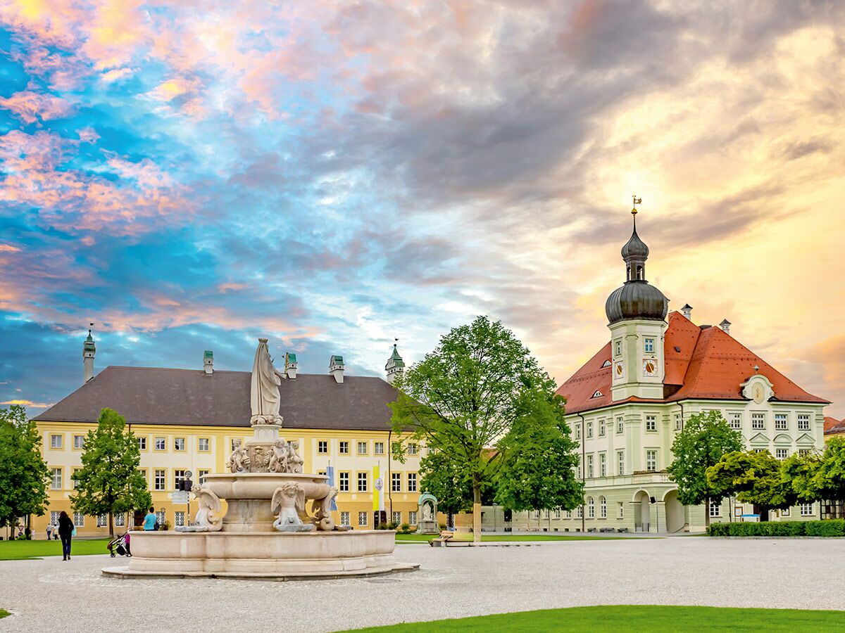 Buntes Abendlicht über Kloster Andechs mit barockem Domturm und detailreichem Brunnen auf dem Vorplatz, Bayern.
