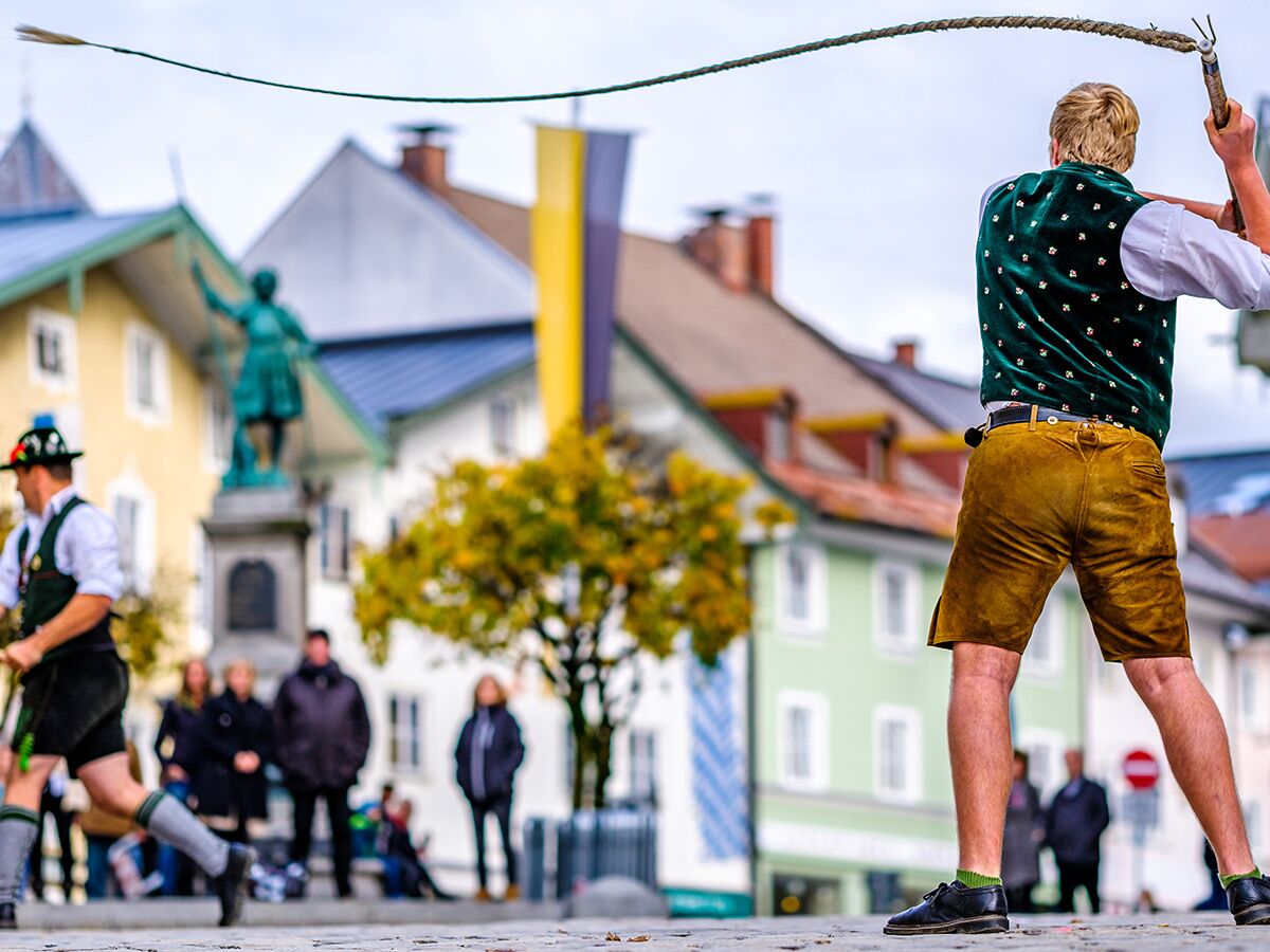 Mann in Lederhose beim traditionellen Goaßlschnalzen auf einem Stadtfest in Bayern, bunte Häuser im Hintergrund.