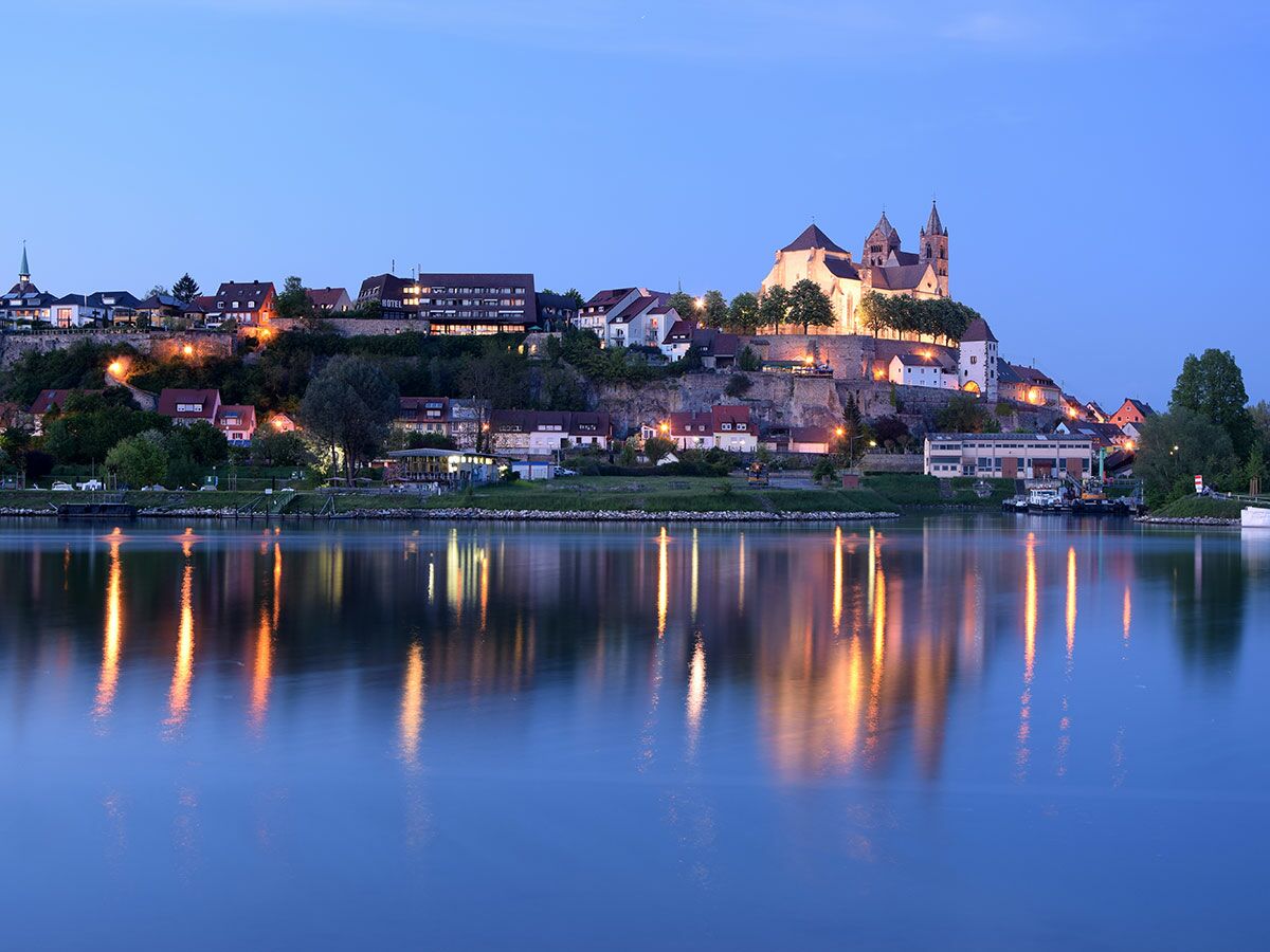 Abendliche Panoramaansicht einer beleuchteten historischen Stadt mit Dom auf einem Hügel, Spiegelung im ruhigen Flusswasser.
