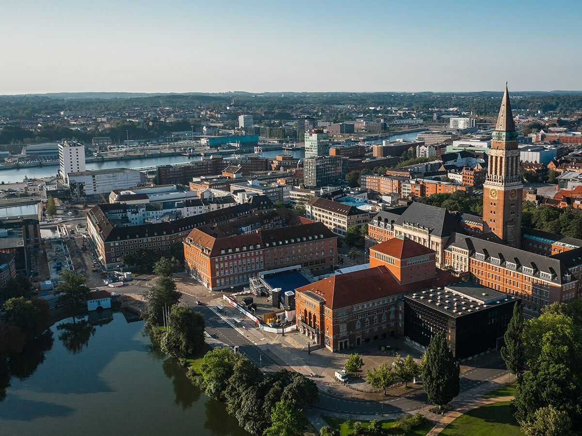 Luftaufnahme des Kieler Rathauses mit seinem ikonischen Turm, Kleiner Kiel und der Kieler Förde bei Sonnenschein.