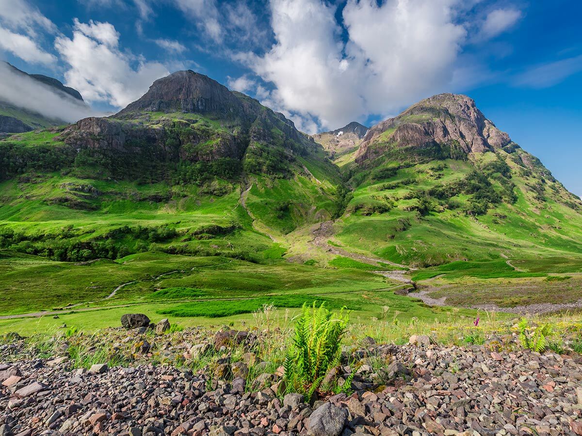 Majestätische grüne Berge in Glencoe, Schottland, mit einem Tal, blauem Himmel, weißen Wolken und felsigem Vordergrund.