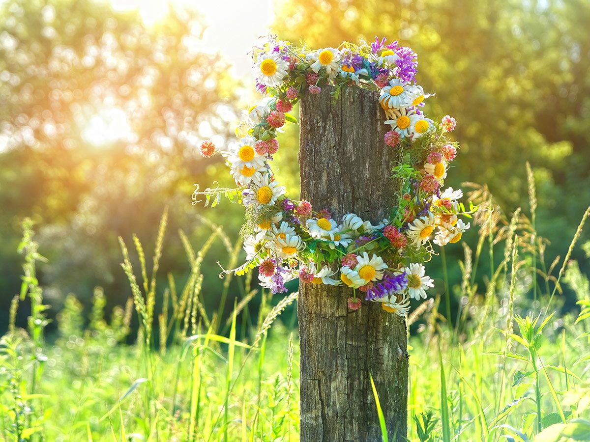 Bunter Blumenkranz aus Margeriten und Kleeblüten hängt an rustikalem Holzpfahl in sonniger Sommerwiese.