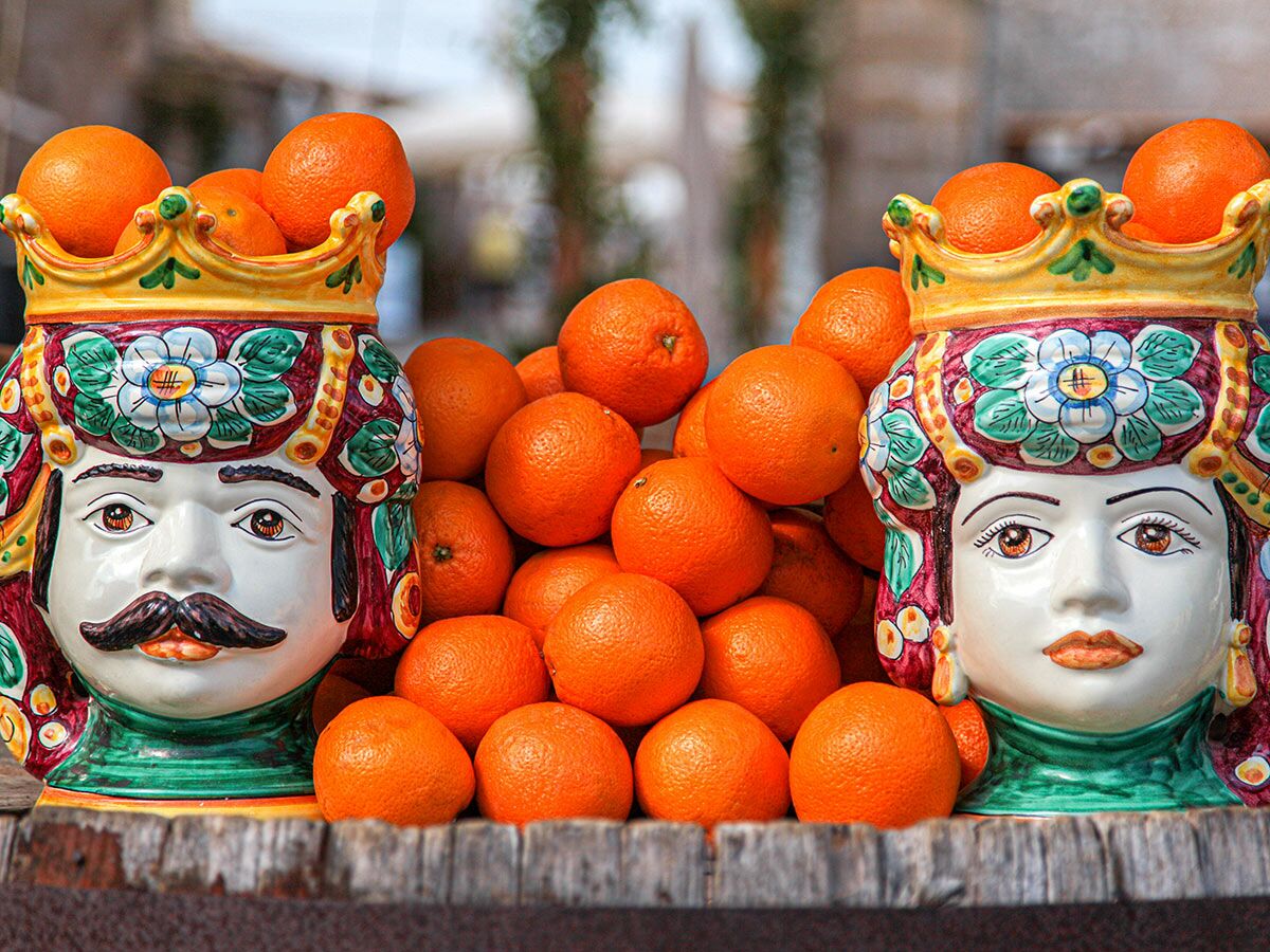 Zwei farbenfrohe sizilianische Keramik-Köpfe (Teste di Moro), gefüllt mit und umgeben von leuchtenden Orangen, auf Holz.
