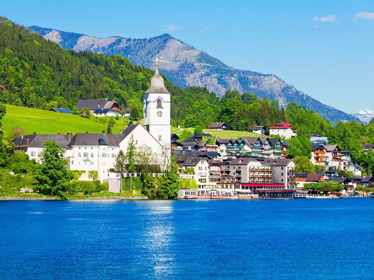 Idyllisches Alpendorf mit weißer Kirche und bunten Häusern am Ufer eines blauen Sees, umgeben von grünen Bergen unter klarem Himmel.