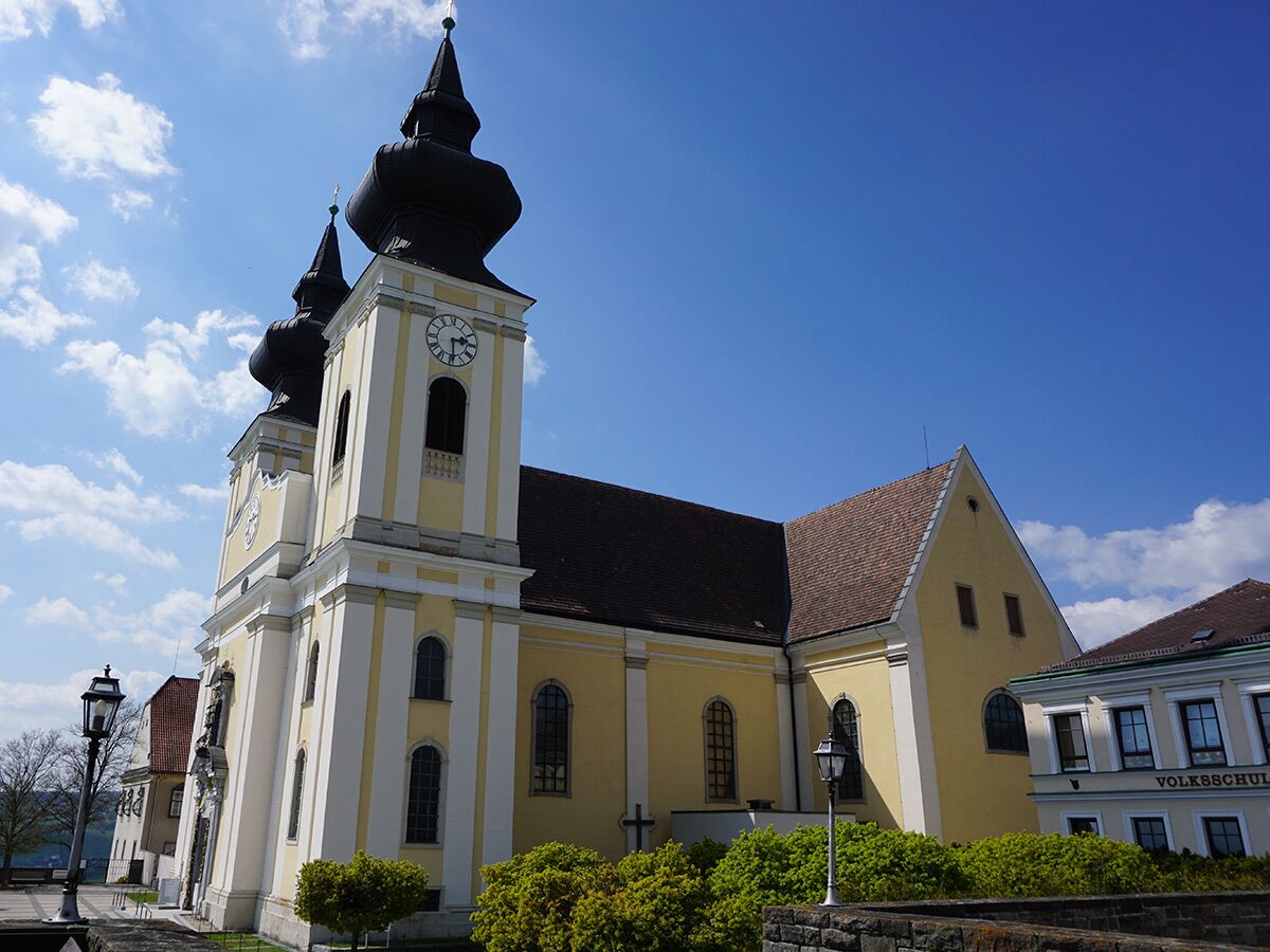 Gelbe Barockkirche mit zwei markanten schwarzen Zwiebeltürmen unter blauem Himmel mit weißen Wolken, grüne Büsche im Vordergrund.