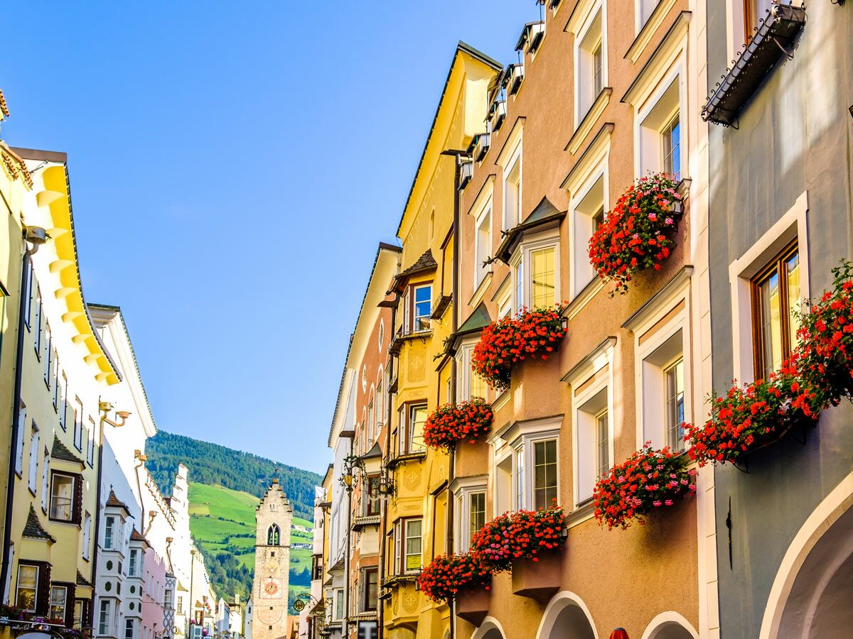 Charmante Altstadtgasse in den Alpen mit farbenfrohen Häusern, roten Geranien in Blumenkästen und Kirchturm im Hintergrund unter blauem Himmel.