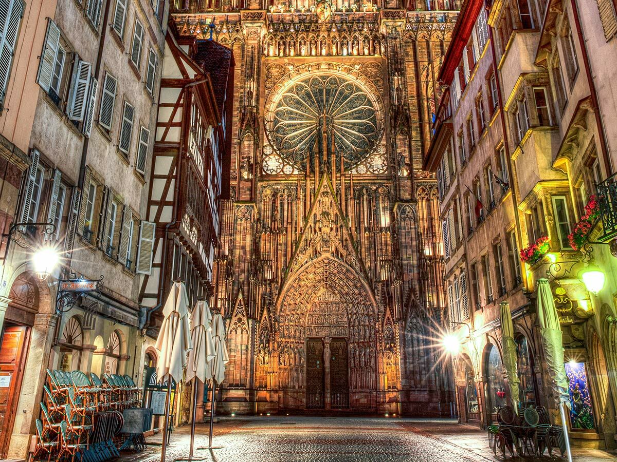 Die beleuchtete Fassade der Straßburger Kathedrale bei Nacht, umgeben von historischen Gebäuden mit Cafés und Kopfsteinpflaster.