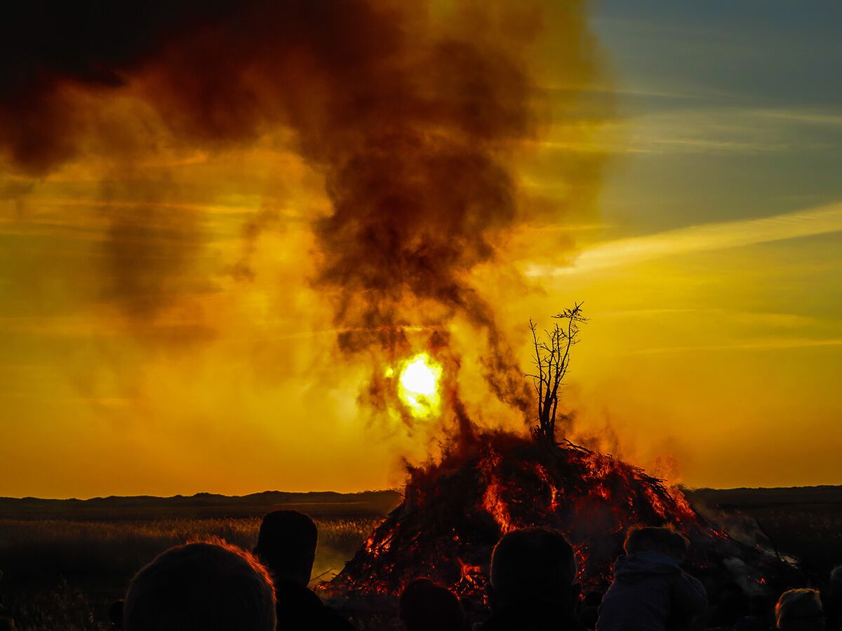 Ein großes Osterfeuer brennt bei Sonnenuntergang und erzeugt dramatischen Rauch, während Menschen zusehen.