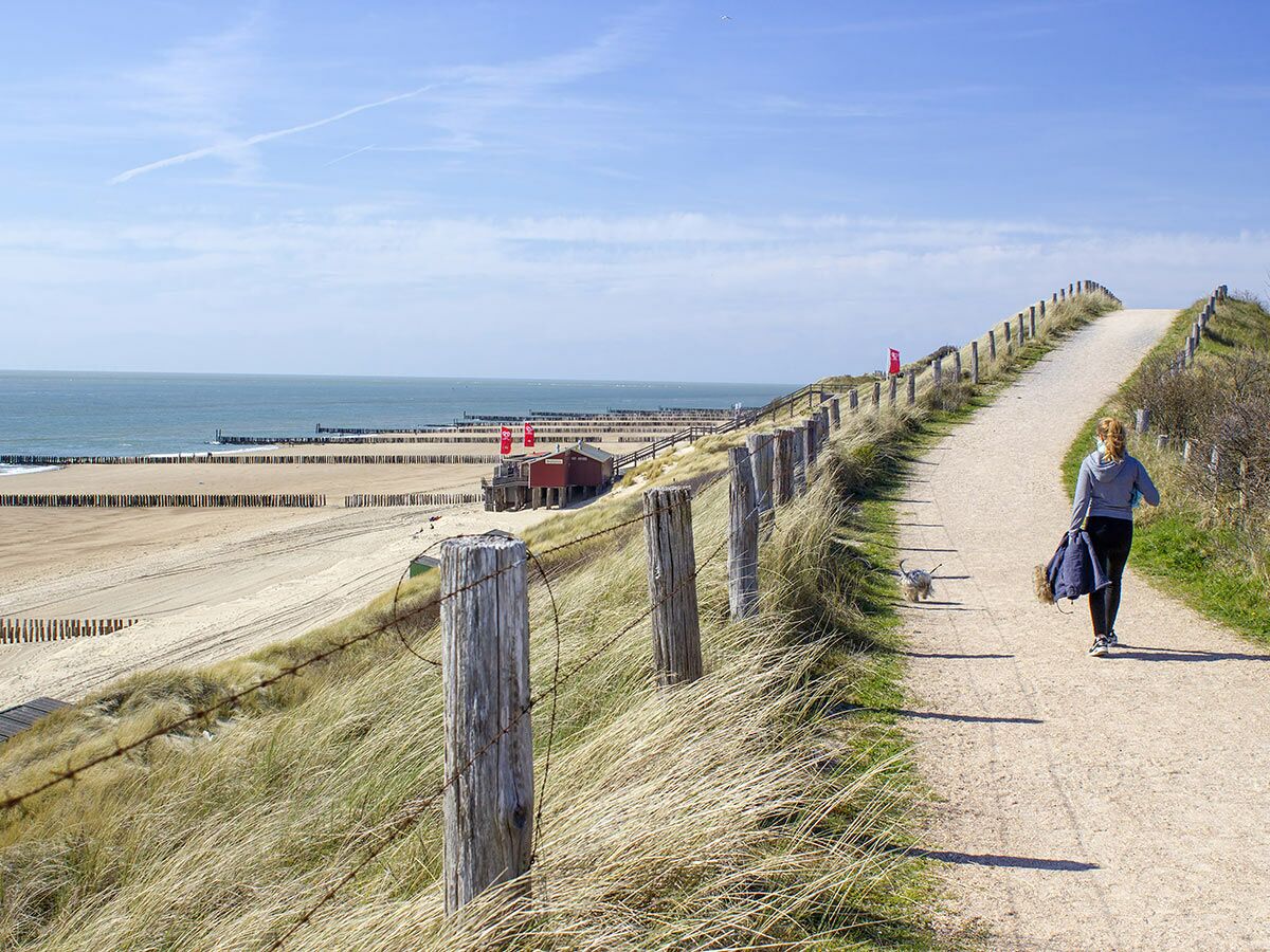 Frau spaziert mit Hund auf Dünenweg am Strand, dahinter Meer mit Buhnen und rotem Gebäude unter blauem Himmel.