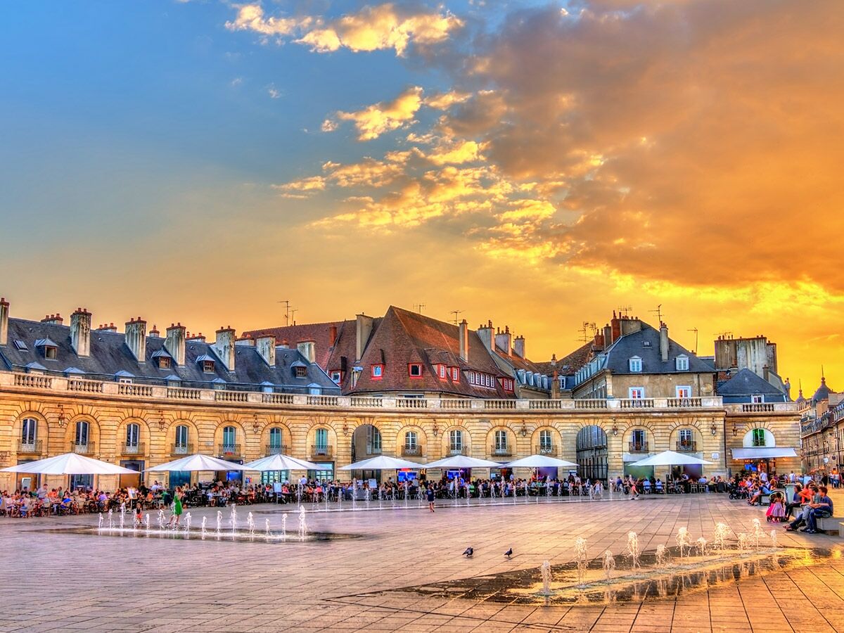 Lebhafter Sonnenuntergang über dem Place de la Libération in Dijon, Frankreich, mit Menschen beim Abendessen an den Springbrunnen.