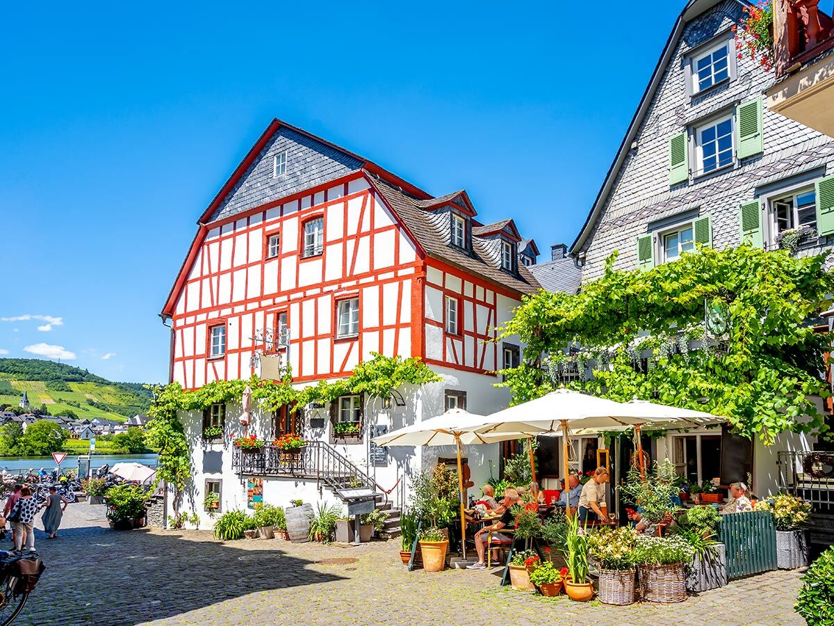 Sonniges Dorf am Fluss mit roten Fachwerk-Häusern, Weinbergen und belebter Café-Terrasse.