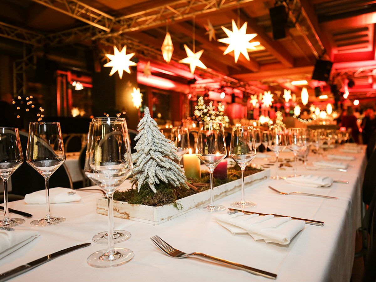Ein festlich gedeckter Tisch in einem Restaurant mit weißem Tischtuch, Gläsern, Besteck und Weihnachtsdekoration mit Baum, Kerzen, Moos. Hintergrund: beleuchtete Sterne.