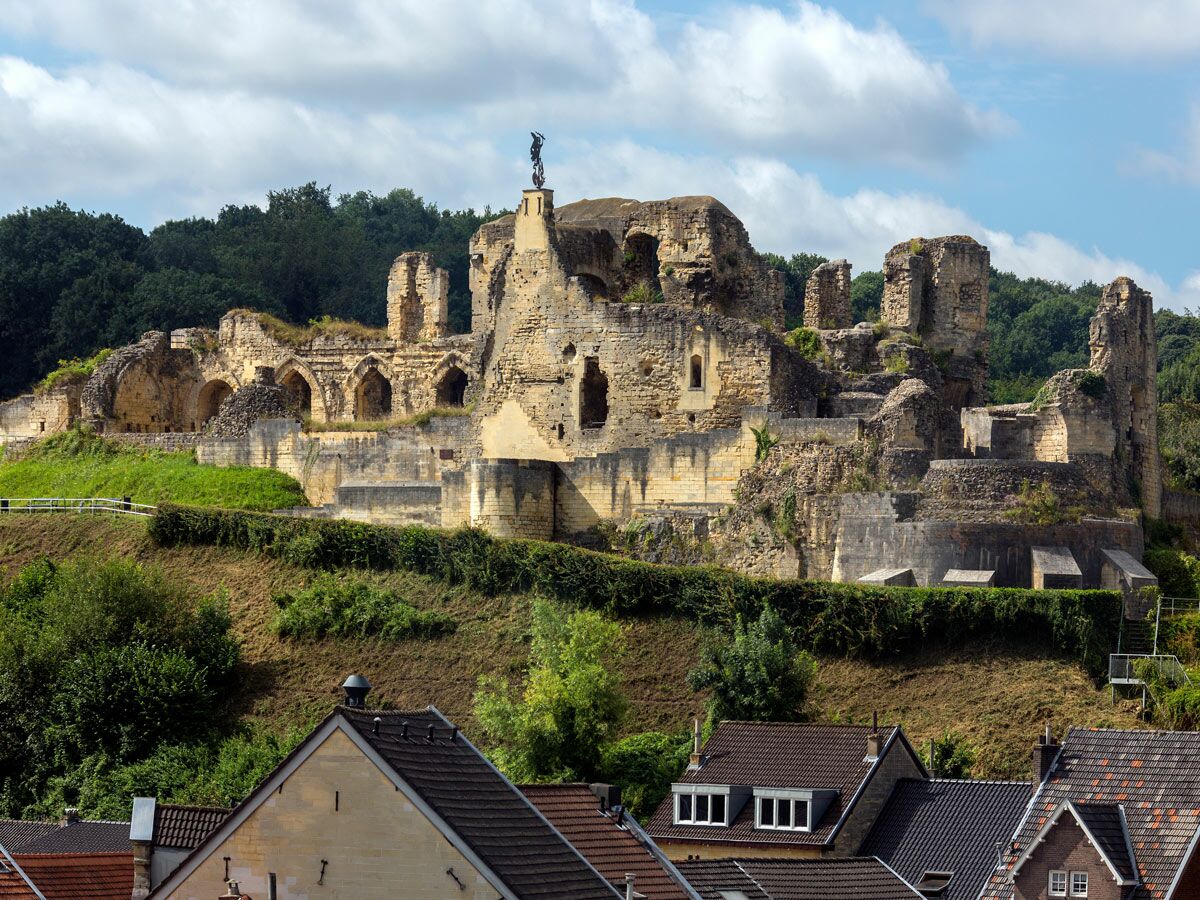 Weitläufige Burgruine Valkenburg in den Niederlanden auf einem grünen Hügel mit Häusern im Vordergrund unter blauem Himmel.