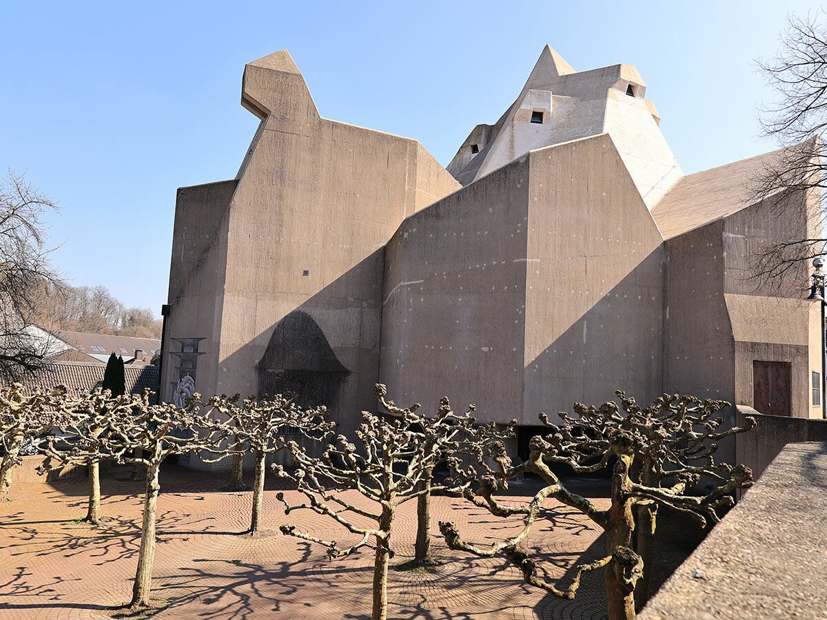 Brutalistische Betonkirche mit scharfen Kanten unter blauem Himmel, davor geometrisch geschnittene Platanen im Winterlicht.