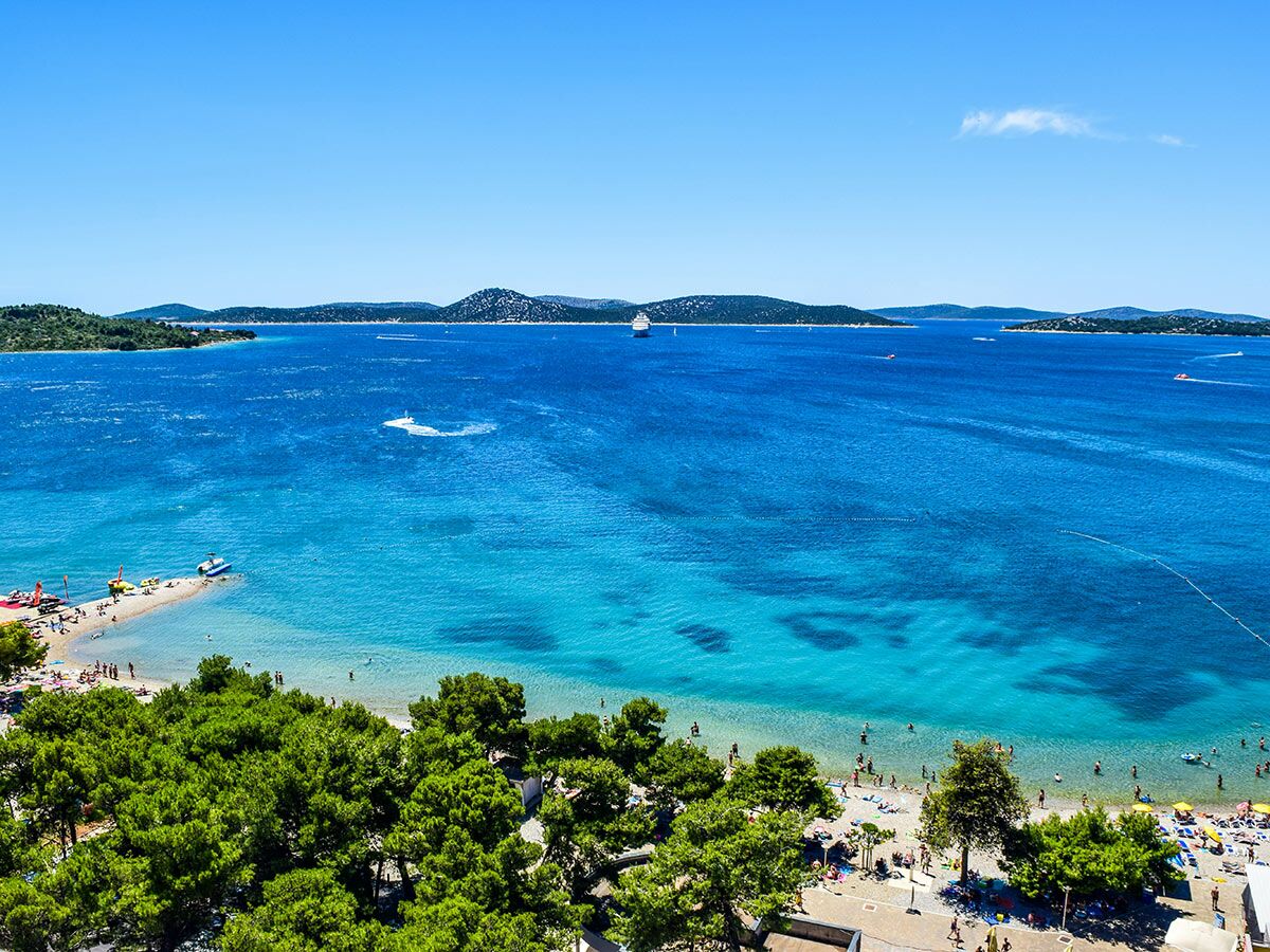 Luftaufnahme eines sonnigen Strandes mit Schwimmern, Booten und Jet-Skis im türkisen Meer vor grünen Inseln.