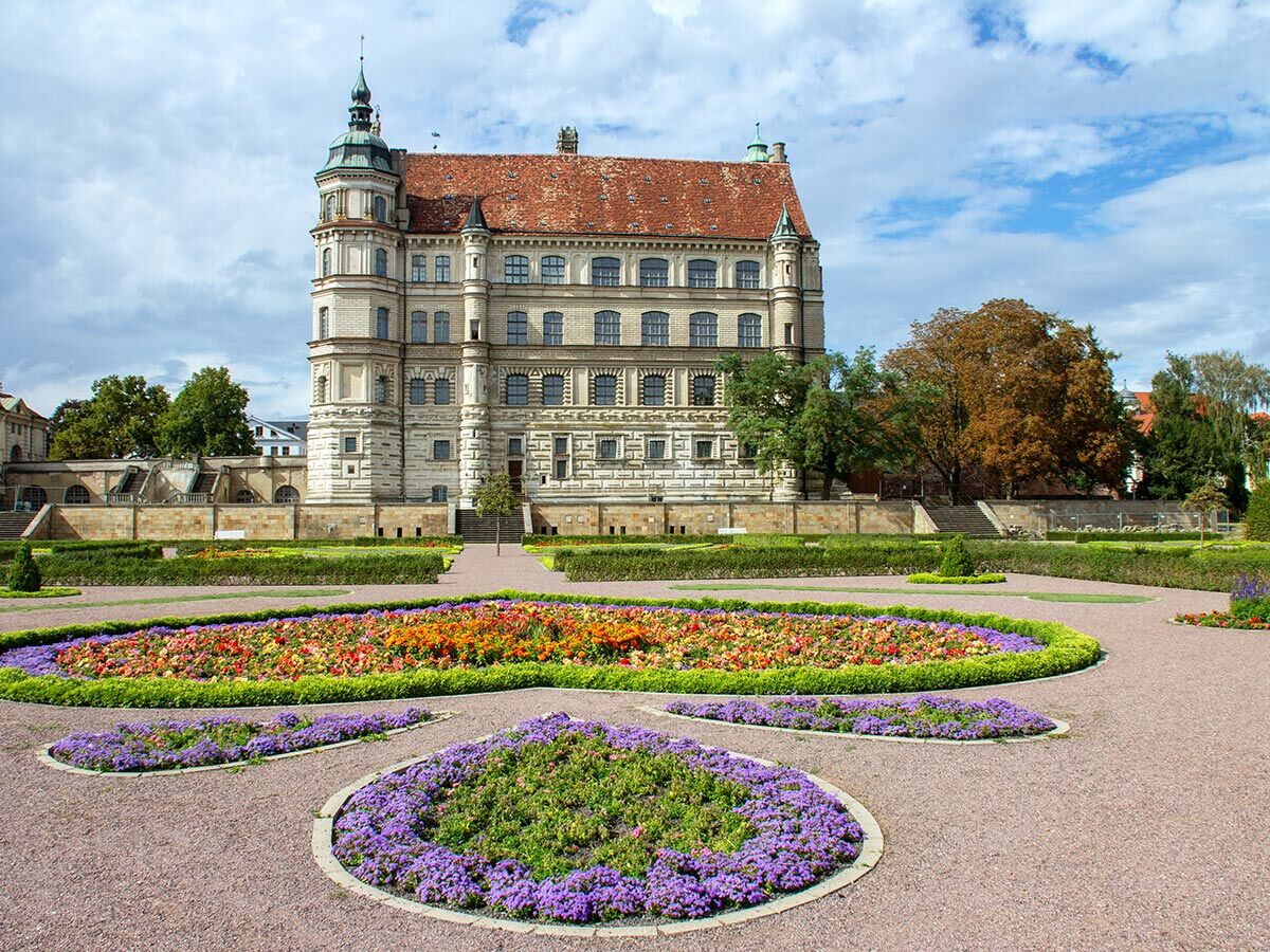 Schloss Güstrow, ein historisches Renaissance-Schloss mit rotem Dach, Turm und formalen Gärten voller bunter Blumen.
